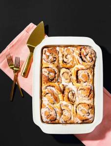 Casserole dish of cinnamon rolls on a pink linen, with gold serving ware on a black background